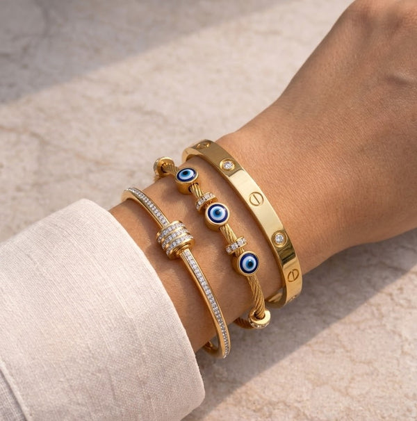 "Woman's wrist wearing a trendy 3-piece 18k gold bracelet stack featuring an evil eye charm bangle, a classic screw motif cuff, and a crystal roller bracelet."