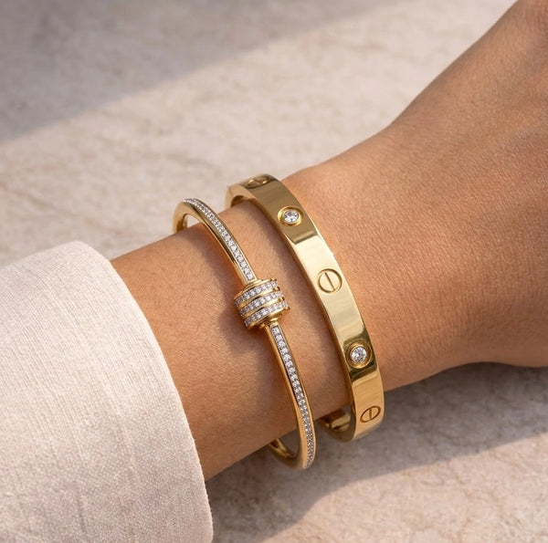 A close-up view of a woman's wrist wearing the complete Luxe Gold Bangle Set, showcasing the delicate split pavé barrel bangle stacked with the iconic screw-motif bangle on a neutral, sunlit stone surface.