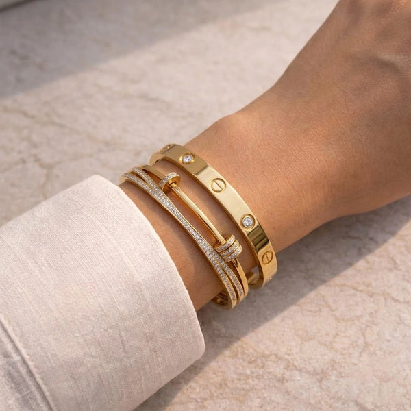 A polished, curated stack of a classic gold love bangle with screws and a modern layered knot bracelet with cz diamonds, resting on white silk fabric.