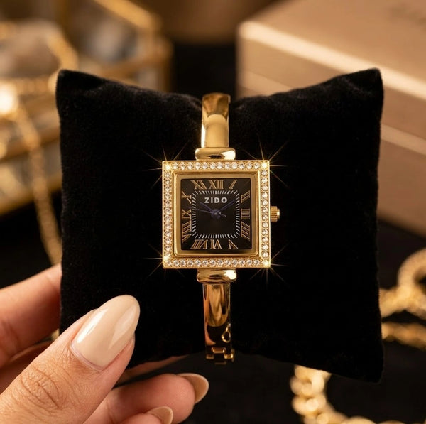 Close up of square dial gold plated watch with studded bezel resting on a black velvet cushion.